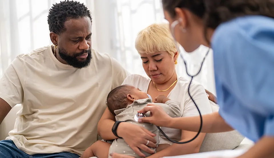 Parents holding their infant child while a caregiver uses a stethoscope.