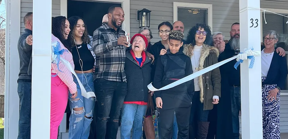 A diverse group of 14 people stand on the front porch of a home as the ribbon-cutting ceremony proceeds.