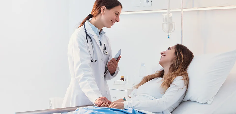 A female doctor standing next to a patient's bed. The doctor is smiling at the female patient, who is also smiling at the doctor.
