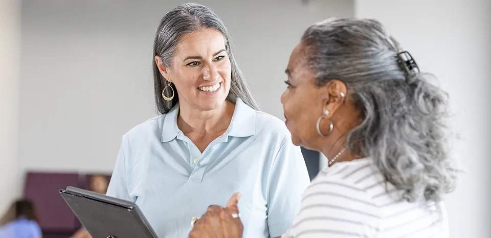 A woman with a tablet speaks to another woman in a hospital setting.