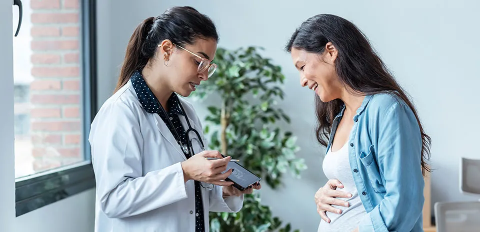 A doctor on the left showing a patient on the right a picture of an ultrasound. The patient on the right holds her stomach, smiling at the ultrasound.