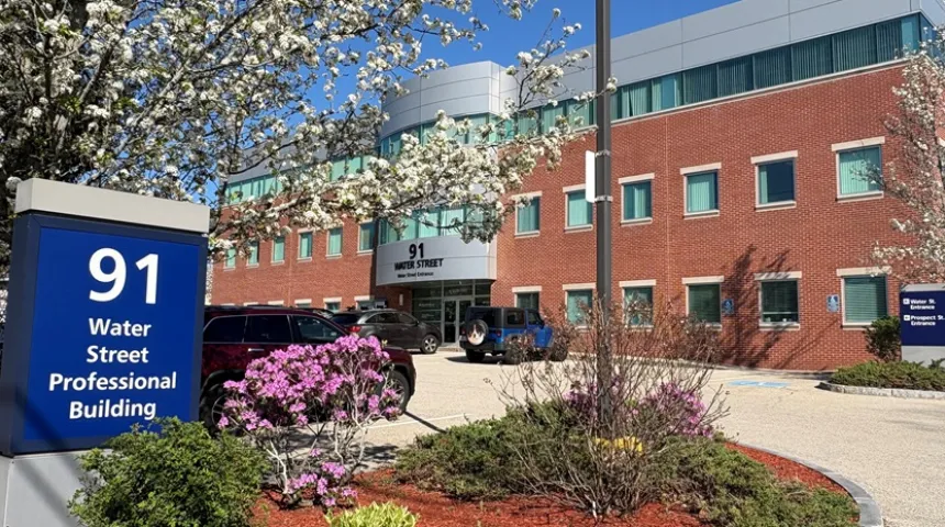The exterior of the brick building at 91 Water Street in Milford, Mass. on a sunny day. Flowers are blooming on trees.
