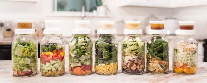 A set of mason jar salads with containers of dressing have been assembled on a countertop.