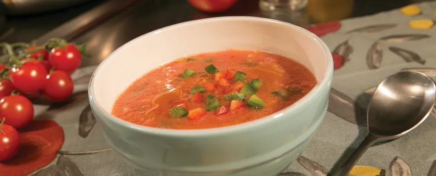 A bowl of gazpacho is is sitting next to a spoon and cherry tomatoes.