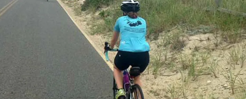 A woman in a blue shirt is riding a bike along a sandy road.